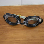 Black and clear swimming goggles with a beige strap resting on a light brown wooden surface.