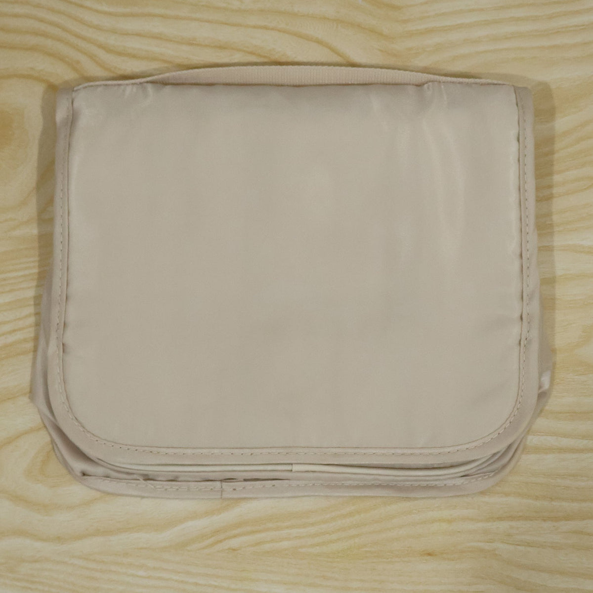 A neatly folded beige toiletry bag with a subtle sheen, resting on a light wooden background.