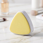 A white triangular makeup sponge holder with a yellow sponge sits on a marble surface with blurred cosmetics in the