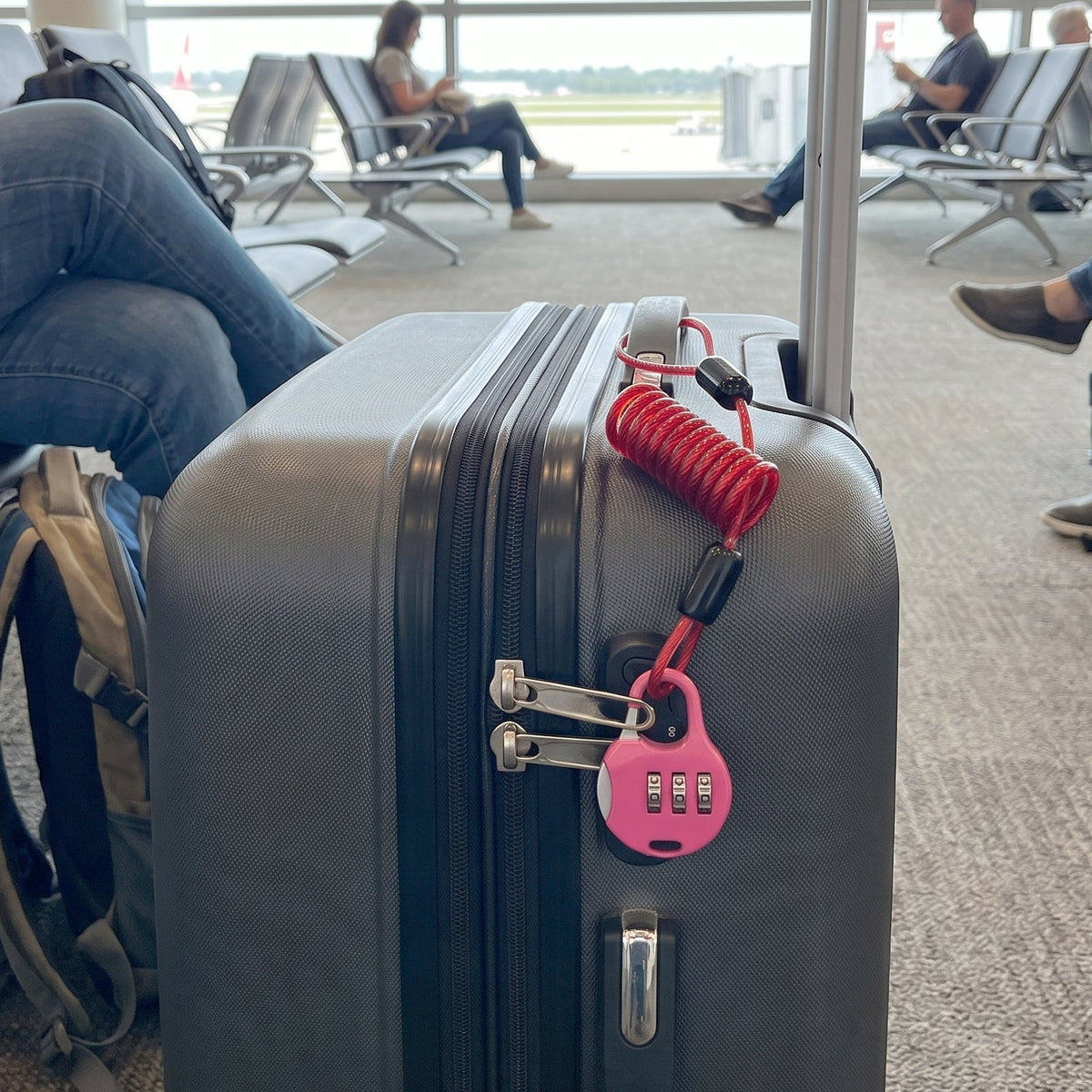 A pink combination lock with a red coiled cable secures a gray suitcase at an airport gate.