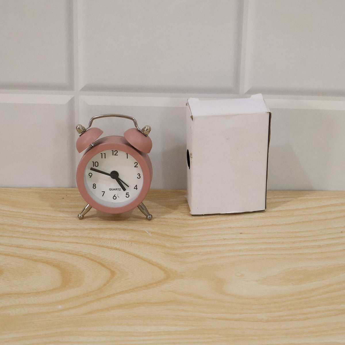 Pink twin bell alarm clock next to a small white open box on a light wooden surface with white tiled wall.