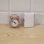 Pink twin bell alarm clock next to a small white open box on a light wooden surface with white tiled wall.