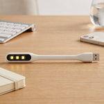 A white flexible USB LED light with three yellow LED bulbs sits on a light wooden desk next to a notebook and phone.