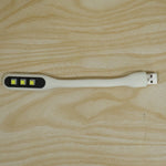 A white flexible USB LED light with three yellow LED bulbs and a USB-A connector resting on a light wood grain surface.
