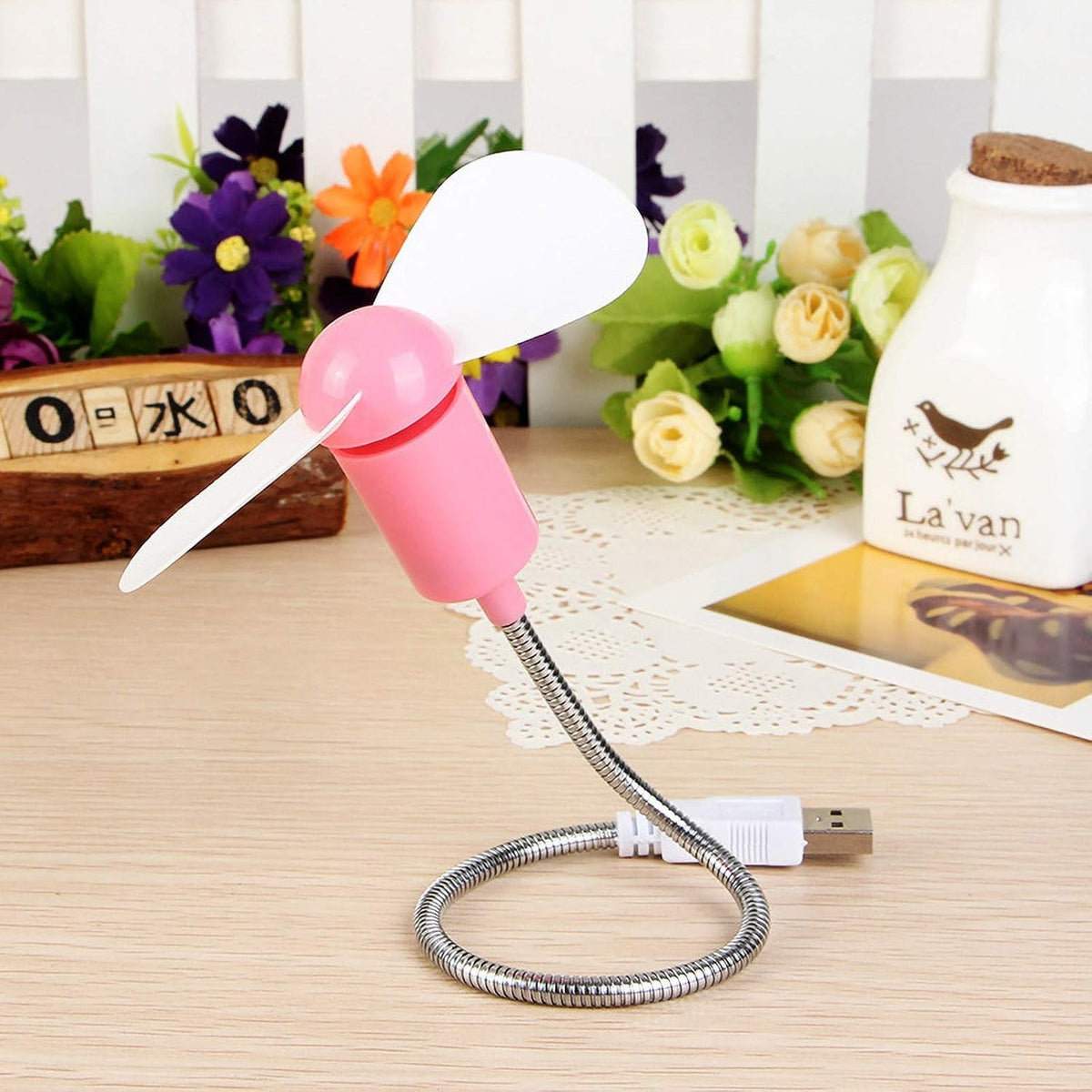 Pink USB mini fan with white blades on a wooden desk, surrounded by decorative flowers and a ceramic bottle.