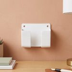 A white wall-mounted phone holder with two compartments on a peach-colored wall above a wooden desk.