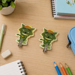 Two green cartoon goblin-like characters with hats, acting as wall hooks, on a wooden desk with stationery.