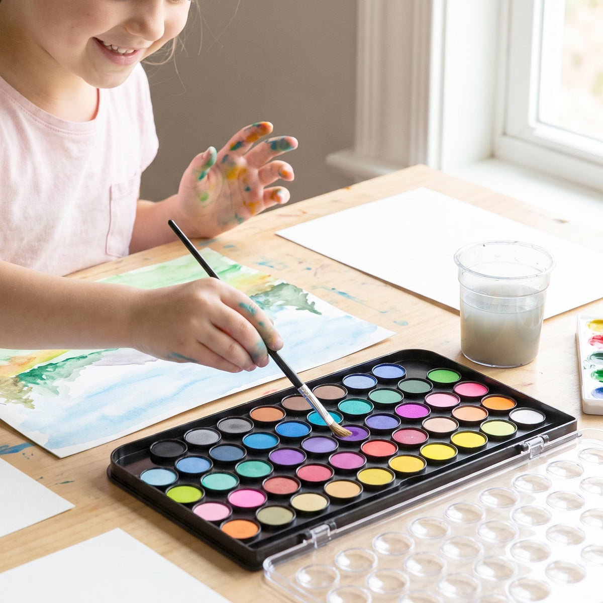 A child's hands, stained with paint, using a brush to paint with a 48-color watercolor set on a wooden table.
