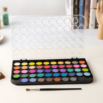 An open black watercolor palette with 48 vibrant solid colors and a clear lid, with a paintbrush and books in the background.