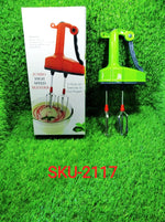 A green manual hand blender with whisks sits on artificial grass next to its white product box, which features an orange