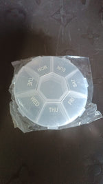 A clear 7-day pill organizer, still in its plastic packaging, on a dark surface.