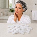 A woman in a white robe wearing a clear shower cap, with many folded shower caps on a counter below.