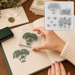 A hand presses a clear rubber stamp of a tree and birdhouse onto a notebook page, leaving a green ink impression.