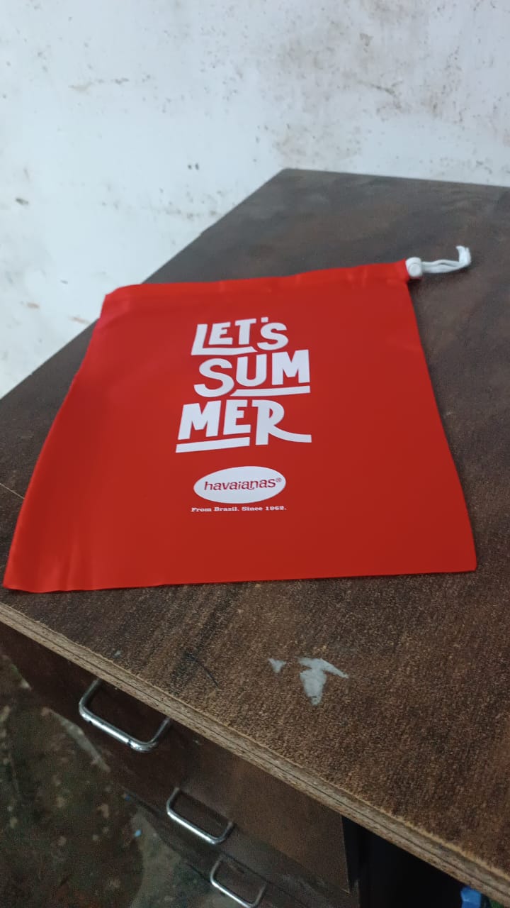 A red drawstring bag with "LET'S SUMMER" and Havaianas logo, resting on a dark wooden desk with metal handles.