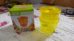A yellow plastic manual food grinder and its product box are on a patterned surface, with various items blurred in the