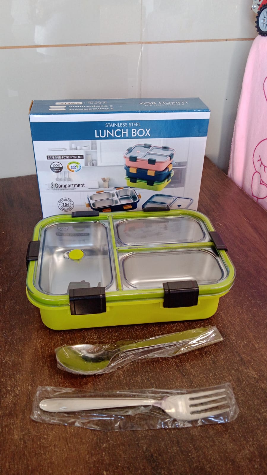 A green three-compartment stainless steel lunch box with dark clips, a spoon, fork, and its packaging box.