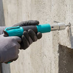 A person wearing gray gloves operates a teal and black manual nail gun, driving a nail into a concrete wall.