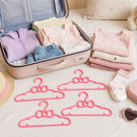 Four pink bow-shaped plastic hangers lie on a bed next to an open suitcase filled with baby clothes.