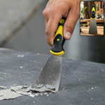 A hand uses a black and yellow handled putty knife to scrape paint chips from a dark surface.