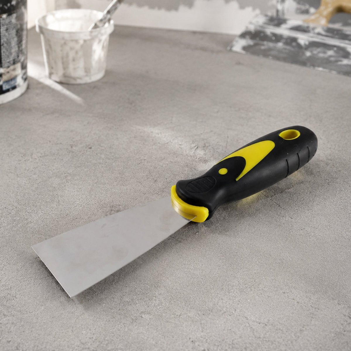 A putty knife with a black and yellow handle lies on a light gray concrete surface, with paint supplies in the background.