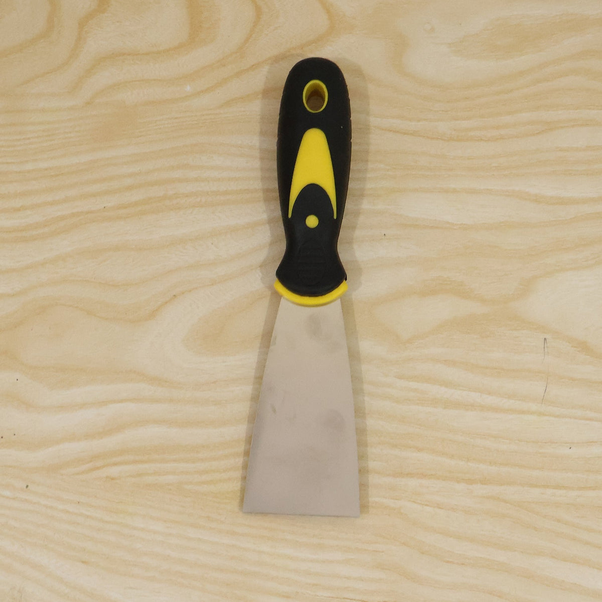A stainless steel putty knife with a black and yellow handle is centered on a light wooden background.