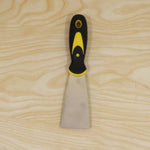 A stainless steel putty knife with a black and yellow handle is centered on a light wooden background.