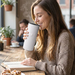 A woman sips from a white thermal tumbler with a heart detail, steam rising, while reading a book in a cafe.