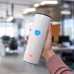 A hand holds a white thermal tumbler with a glowing blue heart and a red temperature display in an office.