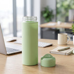 A light green glass water bottle with a silicone sleeve and its detached cap sitting on a wooden desk with a laptop and