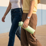 Two people walking, one holding a light green glass water bottle with a silicone sleeve in their hand.