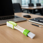 A white and green 3-in-1 cleaning brush resting on a wooden desk with a laptop and office supplies.