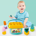 A smiling baby sits behind a blue and yellow shape sorter bucket with various colorful blocks around it on a light blue