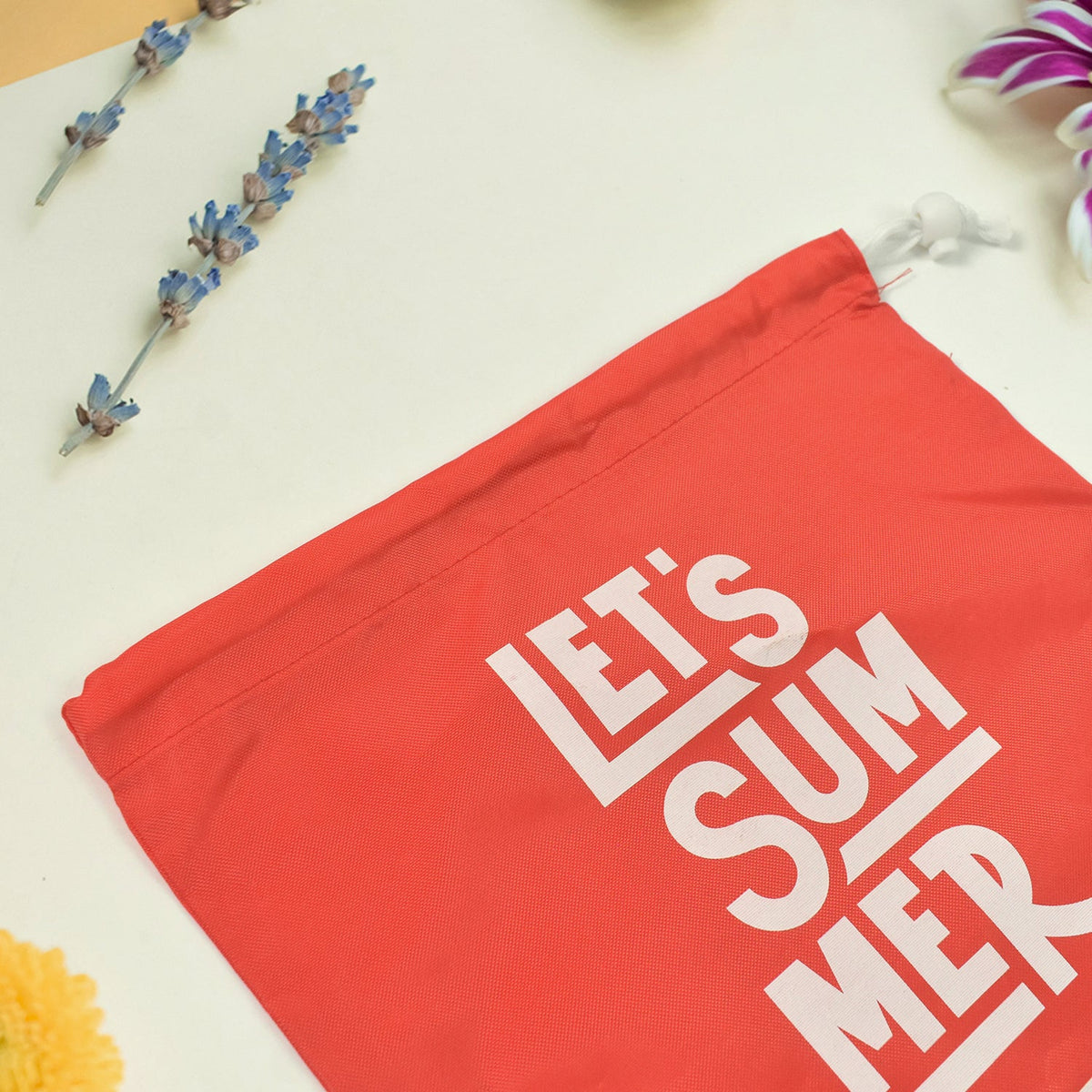 Close-up of a red drawstring bag with "LET'S SUMMER" and Havaianas logo, with purple and yellow flowers in the background.