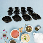 A black ceramic tea set with eight cups and four pineapple-shaped snack bowls on a blue surface, surrounded by tea leaves