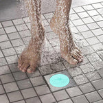 A light blue silicone drain strainer is on a tiled shower floor as water splashes around bare feet.