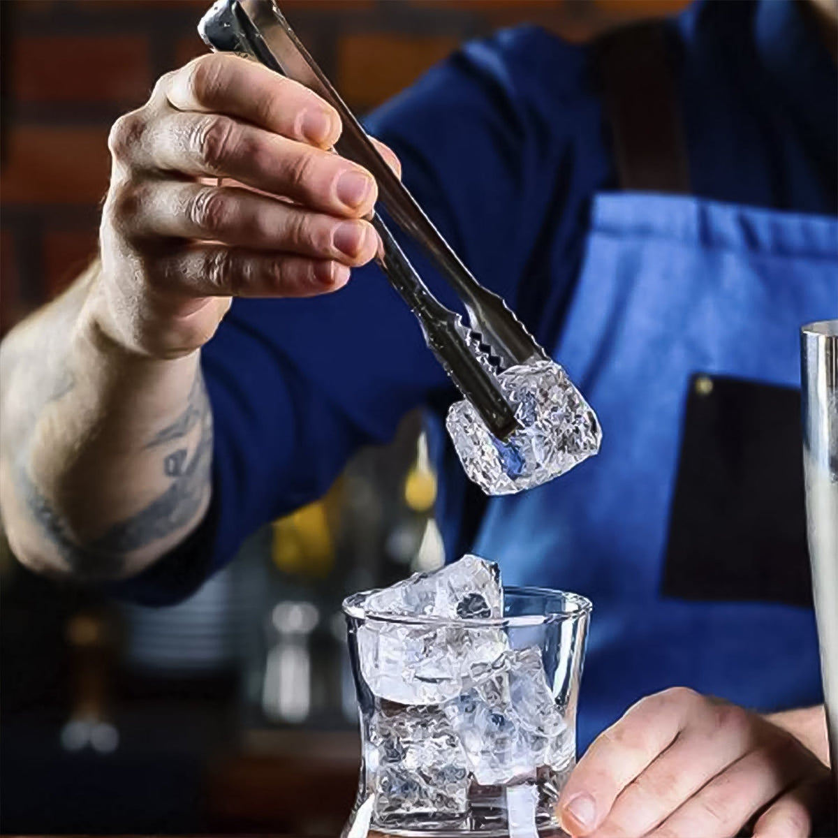 A person in a blue apron uses a silver tong to place an ice cube into a glass on a bar counter.