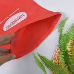 A hand opening a red drawstring bag with the Havaianas logo, next to a woven mat and green fern leaves.