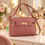 A mauve sling bag with a gold bow and colorful charms sits on a vanity table with flowers and perfume bottles.