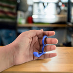 A hand demonstrates holding a blue plastic finger ring phone holder with two fingers.