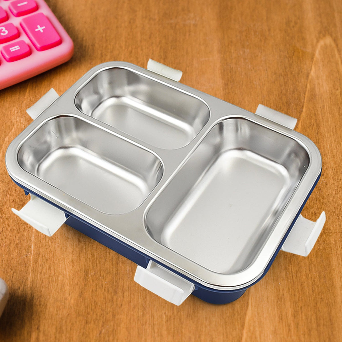A blue lunch box with three stainless steel compartments and white clasps, with a spoon, in front of its product box.