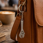 A silver metal mini adjustable wrench keychain hangs from a brown leather bag strap, with a coffee cup in the background.