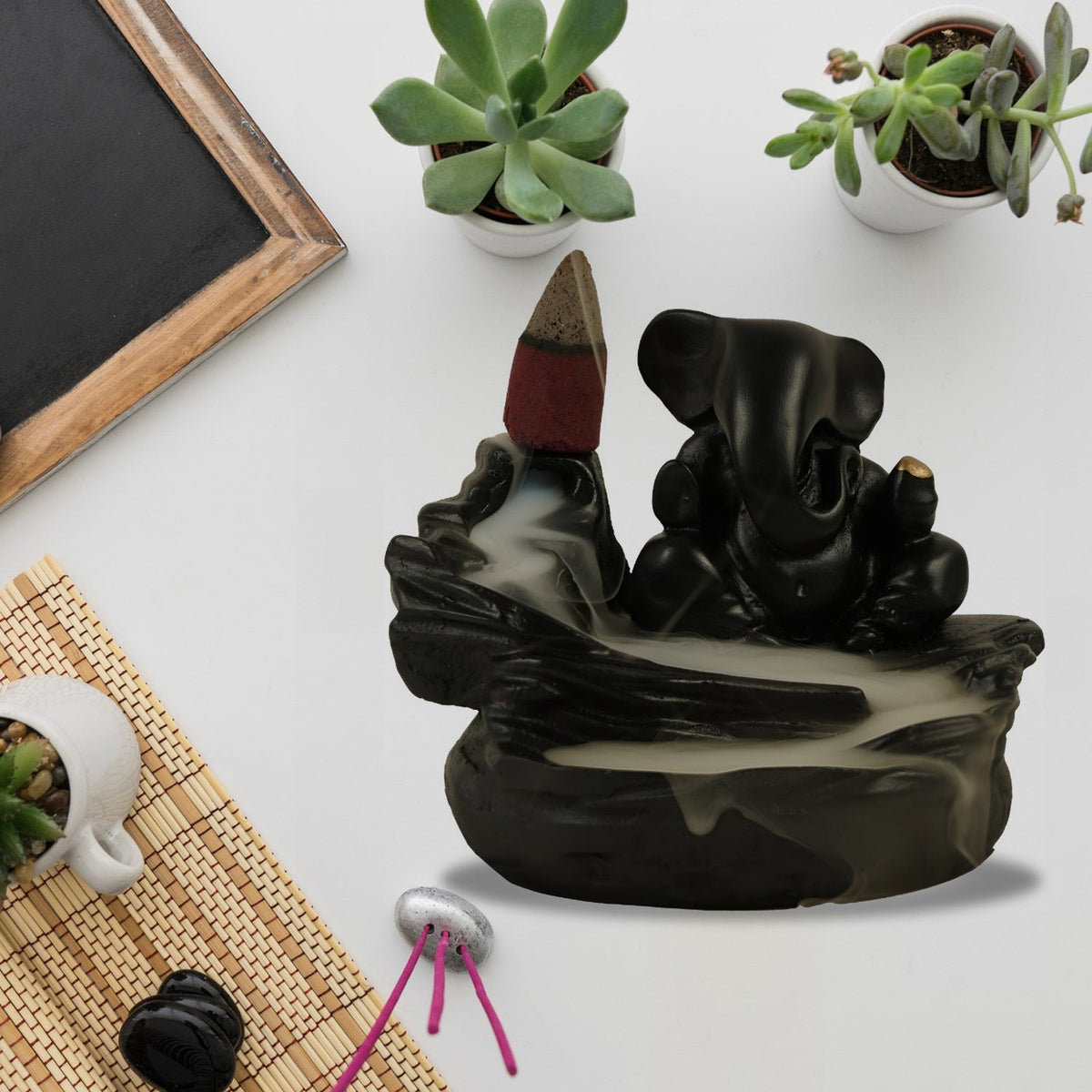 A black Ganesha backflow incense burner with smoke, succulents, and a small mat on a white surface.