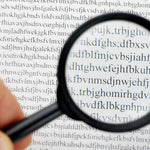 A hand holds a black magnifying glass over a page of text, enlarging the letters for easier reading.