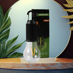 A black and clear portable LED tent light with a black carabiner hangs in front of its green and black packaging, set
