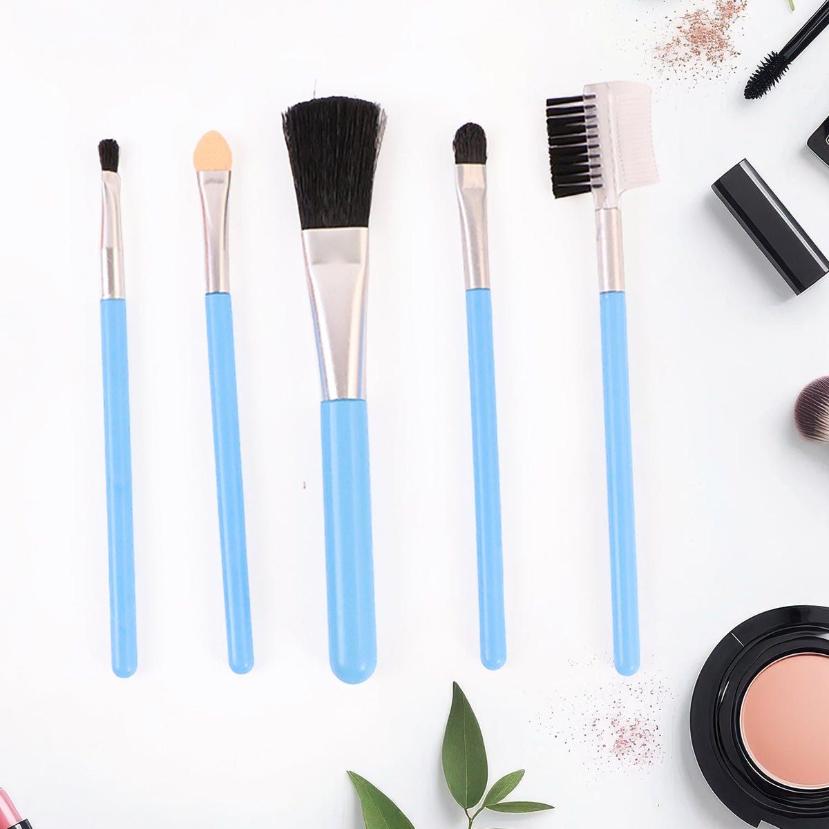 Five light blue makeup brushes are arranged on a white background, with scattered makeup powder and green leaves.