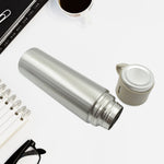 The silver vacuum flask lies on a white desk next to its detached beige cap, with a notebook and glasses nearby.