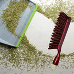 A red and white handled brush with brown bristles lies next to a gray dustpan, surrounded by green dried herbs.
