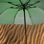 The underside of an open green umbrella reveals its black frame and silver lining, above a textured brown surface.