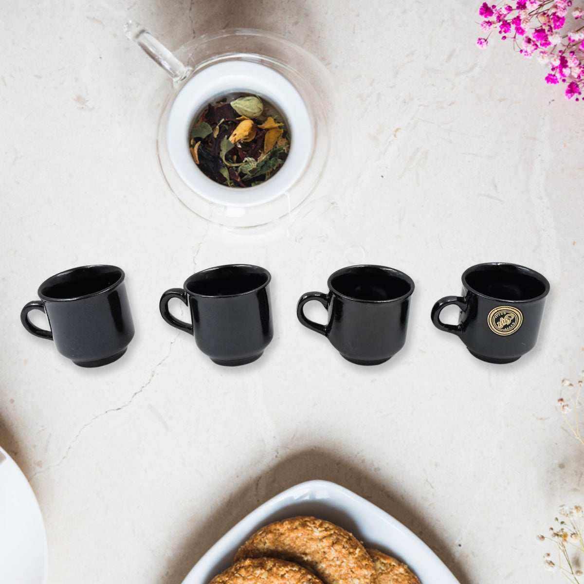 Four black ceramic tea cups with gold accents on a white marble surface next to a glass infuser filled with loose leaf tea.