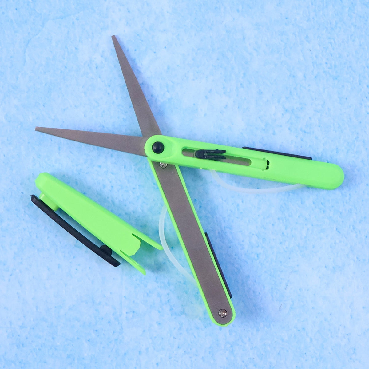 Green pen-shaped scissors with a black safety cutter are displayed on a light blue textured surface.